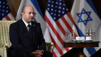 L'image montre un homme assis sur une chaise ornée, vêtu d'un costume sombre. Il a une expression sérieuse et semble concentré. À l'arrière-plan, on peut voir plusieurs drapeaux, dont ceux des États-Unis et d'Israël. Sur la table devant lui, il y a des bouteilles d'eau et des tasses. L'ambiance semble officielle et solennelle.