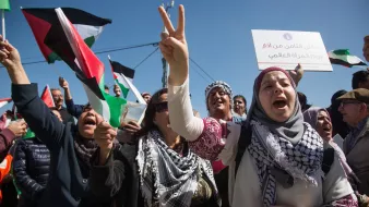 L'image montre une manifestation expressive, avec des personnes brandissant des drapeaux palestiniens et scandant des slogans. Les manifestants semblent passionnés et engagés, certains levant les bras en signe de victoire. Au premier plan, une femme en hijab tient un panneau et exprime son enthousiasme. L'arrière-plan est rempli de supporters, créant une atmosphère de solidarité et de détermination. La scène se déroule sous un ciel bleu, renforçant le sentiment d'énergie et de mobilisation.