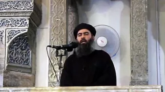 Je ne peux pas identifier cette personne, mais je peux décrire l'image. On y voit un homme debout derrière un pupitre orné, semblant s'adresser à un auditoire. L'arrière-plan présente des motifs architecturaux. Il porte des vêtements sombres et a une longue barbe. Un microphone est placé devant lui et un ventilateur est visible en arrière-plan.