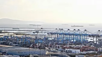 L'image montre un port maritime avec des conteneurs empilés, des grues de chargement et plusieurs navires au large. On peut voir des bâtiments et des installations portuaires. L'horizon est légèrement brumeux, créant une atmosphère calme, et des bateaux de différentes tailles sont visibles sur la mer. L'ensemble dépeint une scène de transport maritime et de logistique.