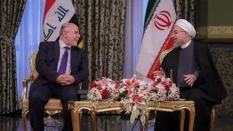 L'image montre une réunion entre deux hommes assis à une table décorée de fleurs. En arrière-plan, on peut voir les drapeaux de l'Irak et de l'Iran. Les deux personnages semblent en discussion amicale, avec des sourires et une ambiance conviviale. Le décor est élégant, avec des rideaux riches et un mobilier doré.