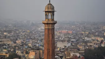 L'image montre un paysage urbain avec une tour élancée qui se dresse au milieu de nombreux bâtiments. La tour a une architecture distinctive, avec des motifs ornés et un dôme en haut. En arrière-plan, on peut voir une vaste étendue de maisons et de structures, témoignant d'une villeDense. L'atmosphère semble légèrement brumeuse, suggérant une pollution ou une humidité ambiante. Les couleurs des bâtiments varient, ajoutant une touche de diversité à l'ensemble de la scène.