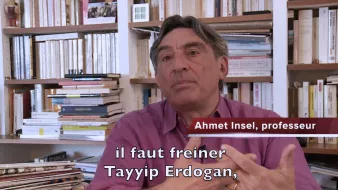 L'image montre un homme assis dans un environnement intérieur, entouré de livres et de dossiers sur des étagères. Il semble s'adresser à un interlocuteur ou à une caméra. L'homme est identifié comme Ahmet Insel, un professeur, et il exprime une opinion sur la nécessité de freiner Tayyip Erdogan. Il porte une chemise de couleur mauve et utilise des gestes pour appuyer ses propos. Les livres en arrière-plan suggèrent un cadre intellectuel.