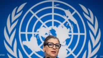 L'image montre une femme parlant devant un microphone, avec un fond représentant le logo des Nations Unies. Elle porte des lunettes et a les cheveux attachés. Son expression semble concentrée, et elle est probablement en train de faire une déclaration ou de répondre à des questions. Le décor bleu est typique des conférences de presse internationales.