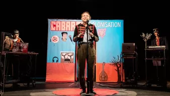L'image montre une scène de spectacle, probablement un cabaret. Au centre, un artiste souriant est sur scène avec un micro, habillé de manière théâtrale. En arrière-plan, un grand panneau coloré affiche le mot "CABARET" et "COLONISATION", suggérant un thème spécifique. De chaque côté, on aperçoit des musiciens ou des techniciens avec des instruments ou des équipements, contribuant à l'atmosphère festive et engageante. Le sol est orné d'un tapis rouge, ajoutant une touche de mise en scène au spectacle.