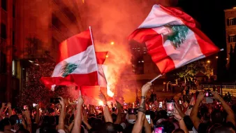 L'image montre une foule rassemblée, brandissant des drapeaux du Liban, dans une ambiance festive et animée. On peut voir des fumigènes rouges qui créent une atmosphère vibrante. De nombreuses personnes semblent capturer ce moment avec leurs téléphones. La scène se déroule probablement la nuit, avec des lumières urbaines en arrière-plan. L'engouement et la camaraderie parmi les participants sont palpables.