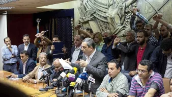 L'image montre une conférence de presse ou une réunion. Un homme se tient au centre, parlant devant un groupe de journalistes, avec plusieurs microphones devant lui. Derrière lui, des personnes semblent exprimer leur soutien ou leur désaccord, levant les poings. L'arrière-plan présente une sculpture en relief qui ajoute de la profondeur à la scène. L'atmosphère semble tendue et engagée.