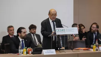 L'image montre un homme âgé, probablement un orateur, se tenant derrière un podium avec un panneau portant le mot "CEDRE" et la date de l'événement. Il semble prendre la parole lors d'une conférence. Autour de lui, plusieurs autres personnes sont assises, attentives, et il y a des bouteilles d'eau et des boissons sur la table. L'ambiance semble formelle et professionnelle, suggérant un événement important.