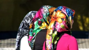 L'image montre le dos de trois femmes portant des foulards colorés. Chaque foulard est orné de motifs fleuris et vibrants, offrant un contraste frappant avec leurs vêtements. Les cheveux des femmes sont couverts, laissant entrevoir seulement leurs silhouettes. L'ambiance semble vivante et pleine de couleurs, soulignant la richesse culturelle des accessoires.