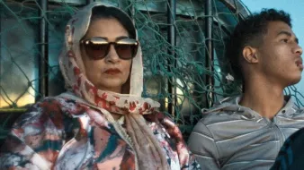 L'image montre une femme et un jeune homme assis ensemble dans un environnement désertique. La femme porte un foulard et des lunettes de soleil, affichant une expression attentive. Elle est vêtue d'une veste aux motifs colorés. À côté d'elle, le jeune homme porte un sweat à capuche et semble pensif, regardant au loin. En arrière-plan, on aperçoit des éléments qui évoquent un paysage aride. L'éclairage donne une atmosphère calme et contemplative à la scène.