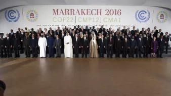 L'image montre un groupe de participants rassemblés lors de la conférence COP22 qui s'est tenue à Marrakech en 2016. Les membres représentés incluent des dirigeants et des délégués de divers pays, vêtus de costumes formels et de tenues traditionnelles. Ils se tiennent debout sur des gradins, souriant pour la photo officielle, avec un fond affichant le nom de l'événement et des logos des Nations Unies. L'ambiance évoque un sentiment de collaboration internationale pour faire face aux défis climatiques.