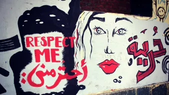 L'image montre une fresque murale qui combine des éléments graphiques et des mots en anglais et en arabe. À gauche, on peut voir le texte "RESPECT ME" en anglais, et en dessous, "احترمني" qui signifie "Respecte-moi" en arabe. À droite, il y a un visage stylisé de femme, avec des yeux expressifs et des lèvres rouges. Le tout est peint sur un mur de couleur claire, créant un message fort sur l'importance du respect.