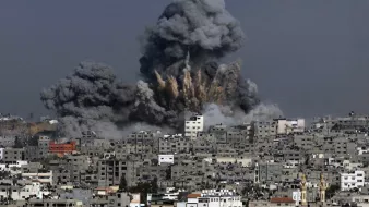 L'image montre une scène de destruction où des colonnes de fumée noire s'élèvent dans le ciel, indiquant qu'une explosion a eu lieu. Les bâtiments sont présents au premier plan, témoignant d'une zone urbaine, et semblent avoir subi des dommages. Le contraste entre la couleur grise de la fumée et les structures environnantes évoque une atmosphère de chaos et de conflit.