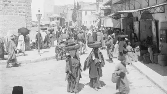 L'image montre une scène animée à la porte de Jaffa à Jérusalem. On y voit de nombreuses personnes en mouvement, portant des vêtements traditionnels. Certains portent des charges sur leur tête, tandis que d'autres se déplacent dans la rue. Des bâtiments historiques se dressent en arrière-plan, et l'atmosphère semble occuper un marché ou un lieu de rencontre. La lumière est vive, ce qui donne une impression de chaleur et d'activité.