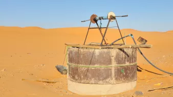 L'image montre une structure située dans un désert de sable doré. Au centre, il y a un puits ou un réservoir en métal, recouvert de tôle, avec un mécanisme en bois au-dessus. Ce dernier semble être utilisé pour pomper de l'eau. La terre environnante est sèche et aride, et on aperçoit des dunes de sable qui s'étendent à perte de vue sous un ciel clair et sans nuages. L'ensemble évoque un environnement désertique, isolé et dépouillé.