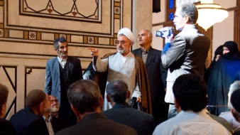 L'image montre un groupe de personnes rassemblées dans un espace intérieur richement décoré. Au centre, un homme en vêtements traditionnels, probablement religieux, semble s'adresser à l'assemblée. Il lève une main en signe d'argumentation ou d'expression. Plusieurs autres hommes l'écoutent attentivement, tandis qu'une ou deux femmes en arrière-plan portent un hijab. L'atmosphère semble sérieuse et respectueuse, avec des détails architecturaux élégants autour.