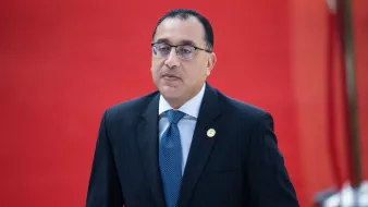 L'image montre un homme en costume noir avec une chemise blanche et une cravate bleue. Il porte des lunettes et a une expression sérieuse. L'arrière-plan est rouge, ce qui crée un contraste vif avec sa tenue. L'homme semble être dans un cadre officiel ou lors d'un événement important.