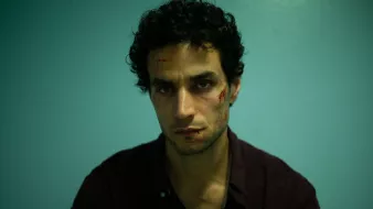 L'image montre un homme assis devant un mur de couleur bleu clair. Il a des cheveux noirs et bouclés, et son expression faciale semble intense ou préoccupée. Son visage présente des éraflures ou des blessures visibles, ce qui suggère qu'il a traversé une situation difficile. Il porte une chemise foncée et a l'air fatigué ou en détresse. L'éclairage est dramatique, mettant en avant ses traits et les blessures sur son visage.