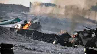 L'image semble représenter une scène de conflit ou de guerre, avec des flammes visibles et de la fumée s'élevant dans l'air. On peut distinguer des structures endommagées et une ambiance chaotique. Le sol est recouvert de débris, et des véhicules militaires ou des équipements peuvent être présents. L'environnement semble aride, suggérant un paysage désertique.
