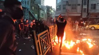 L'image montre une scène dynamique dans une rue urbaine, probablement lors d'une manifestation. On peut voir plusieurs personnes, dont l'une est au premier plan, se tenant près de flammes qui brûlent sur la route. L'atmosphère semble tendue, avec des barrières et des débris éparpillés. La lumière des flammes éclaire les visages des manifestants, qui portent des masques. En arrière-plan, on aperçoit des bâtiments urbains et une lumière tamisée, ce qui donne une impression d'urgence et de mouvement.