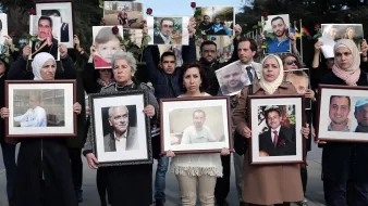 L'image montre un groupe de personnes participant à une manifestation ou un hommage. Elles tiennent des portraits de différents individus, certains avec des roses. Les expressions des participants semblent sérieuses et engagées, suggérant qu'ils rendent hommage à des personnes disparues ou aux victimes d'un conflit. Le décor en arrière-plan semble être en plein air, et plusieurs drapeaux sont visibles. Les vêtements des manifestants varient, avec quelques femmes portant des foulards.