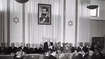 L'image montre une scène à l'intérieur d'une salle de réunion. Au fond, il y a un grand portrait en noir et blanc d'un homme, probablement une figure historique importante, entouré d'étoiles de David. Sur la scène, plusieurs hommes sont assis, en costume, tandis qu'un orateur se tient debout, s'adressant à l'assemblée. L'ambiance semble formelle, indiquant un événement significatif. Les participants, qui sont assis face à la scène, apparaissent attentifs à la présentation en cours.