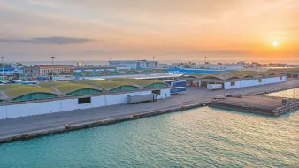 L'image montre un port lors d'un coucher de soleil, avec un paysage industriel. On peut voir des entrepôts aux toits arrondis, bordés par l'eau. Des véhicules et des conteneurs sont visibles, indiquant une activité portuaire. Le ciel est teinté de couleurs chaudes, créant une atmosphère paisible et accueillante.