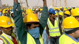 L'image montre un grand groupe de travailleurs, majoritairement en train de lever les bras. Tous portent des casques de sécurité jaunes et des gilets réfléchissants. L'environnement semble être un site de construction, avec plusieurs grues et équipements visibles en arrière-plan. L'atmosphère paraît dynamique et l'affluence des travailleurs indique probablement une activité importante sur le chantier.