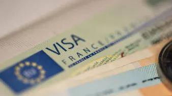 L'image montre un document de type visa, probablement un visa français. On peut voir le mot "VISA" en lettres majuscules, ainsi que des détails graphiques et textuels associés, comme le terme "FRANCE". En arrière-plan, on aperçoit des billets de banque, ce qui suggère un contexte lié à des voyages ou à des transactions. L'ensemble est présenté de manière à mettre l'accent sur le visa.