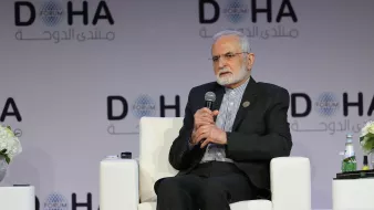 L'image montre un homme assis sur un fauteuil blanc, participant à une conférence ou un forum. Il porte une tenue formelle et semble être en train de s'exprimer ou d'écouter. En arrière-plan, on peut voir un logo avec le mot "DOHA" et d'autres inscriptions en arabe. Des fleurs et une bouteille d'eau sont également visibles sur la scène. L'atmosphère semble professionnelle et tranquille.