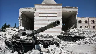 L'image montre un paysage de destruction, probablement lié à un conflit. Au premier plan, on voit des chars endommagés, couverts de débris et de poussière. En arrière-plan, il y a un bâtiment partiellement détruit, dont les murs présentent des marques des destructions. Le ciel est bleu, ce qui contraste avec la scène de désolation au sol. L'ensemble évoque un endroit marqué par la guerre, avec des traces visibles de violence et de désastre.