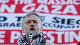 L'image montre un homme prenant la parole devant une grande banderole. Il porte un foulard traditionnel autour du cou et semble s'adresser à une foule. La banderole en arrière-plan contient des messages concernant la situation à Gaza, appelant à mettre fin aux massacres et à la occupation actuelle. L'expression de l'homme indique qu'il est engagé et passionné par le sujet qu'il aborde.