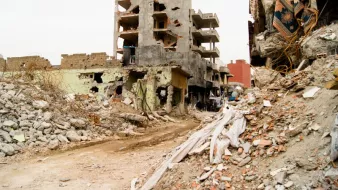 L'image montre un paysage urbain dévasté par la guerre ou une catastrophe. On peut voir des bâtiments partiellement détruits, présentant des murs troués et des structures effondrées. Des débris encombrent le sol, et des morceaux de béton et des matériaux de construction sont éparpillés partout. La rue semble désolée, soulignant l'impact de la destruction sur l'environnement urbain. Les couleurs sont sombres et tristes, renforçant le sentiment de désespoir et de désolation.