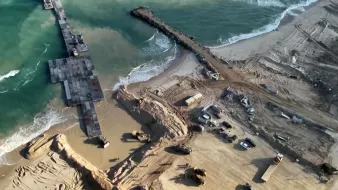 L'image montre une vue aérienne d'un site de construction situé près de la mer. On peut voir un port ou une jetée inachevée s'étendant dans l'eau, entouré de terres travaillées. Des engins de construction sont dispersés autour, et le sol est marqué par des travaux de terrassement. Les vagues de la mer s'échouent à proximité, créant un contraste avec la zone de construction. L'ensemble de la scène illustre une zone en développement, probablement pour une infrastructure côtière.