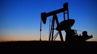 L'image montre un pompage de pétrole silhouetté contre un ciel au crépuscule. On peut voir la structure du pompage avec ses bras articulés, ainsi qu'une échelle menant à la plateforme. Le fond présente des dégradés de bleu et d'orange, indiquant le moment de la journée où le soleil se couche. L'ensemble dégage une ambiance tranquille tout en évoquant l'industrie pétrolière.