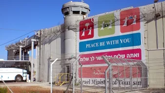 L'image montre un mur imposant, probablement un mur de séparation, avec une tour de surveillance au sommet. À la base du mur, il y a une grande pancarte qui affiche des messages liés à la paix, écrits en plusieurs langues. L'environnement est urbain, et on peut voir un bus stationné à proximité. Le tout est entouré d'une clôture barbelée, ce qui donne une impression de sécurité renforcée. Le ciel est dégagé et ensoleillé.