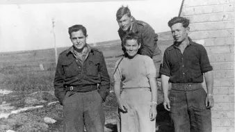 L'image montre quatre personnes se tenant debout à l'extérieur, probablement sur un terrain dégagé. Il y a trois hommes et une femme. Les hommes portent des uniformes militaires ou des vêtements utilitaires, tandis que la femme est habillée simplement dans une robe. L'environnement semble rural, avec une petite bâtisse en arrière-plan et des champs en toile de fond. Tout le monde affiche des expressions neutres ou souriantes, créant une atmosphère de camaraderie.