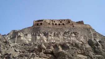 L'image montre une ancienne structure en pierre située sur une colline rocheuse. On peut voir des ruines dissimulées par des murs et des terrasses. Le ciel est dégagé, ce qui donne une belle luminosité à la scène. La végétation est absente, et le paysage est principalement composé de roches. La construction semble avoir une importance historique, suggérant qu'elle pourrait être un vestige d'une civilisation passée.