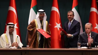 L'image montre quatre hommes debout devant des drapeaux turcs et des Émirats arabes unis. Deux hommes au centre tiennent des dossiers rouges. Les hommes sont vêtus de costumes officiels, et certains portent des habits traditionnels. Ils semblent participer à une cérémonie de signature ou à une conférence officielle. Il y a également des verres d'eau sur la table devant eux. La scène a lieu dans une salle bien éclairée et semble très formelle.