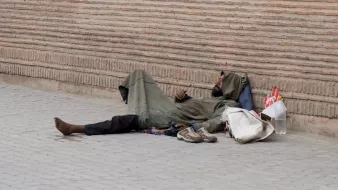 L'image montre une personne allongée sur le sol, semblant sans abri. Elle porte des vêtements sombres et usés, et se trouve à côté d'une paroi de briques. On voit des objets éparpillés autour d'elle, comme des chaussures et un sac. L'environnement semble urbain et le ton de l'image est contemplatif.