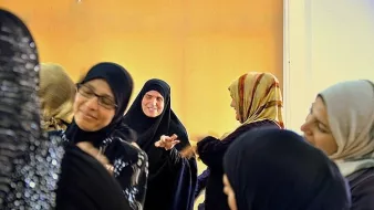 L'image montre un groupe de femmes en discussion ou en interaction. Elles portent des vêtements traditionnels, y compris des hijabs et des abayas. Certaines femmes sourient et semblent engagées dans une conversation joyeuse. L'éclairage dans la pièce donne une ambiance chaleureuse, avec un fond qui semble jaune. L'atmosphère générale est conviviale et animée.