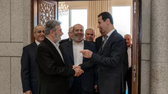 L'image montre un groupe d'hommes en train de se rencontrer dans un intérieur. Plusieurs d'entre eux sourient et semblent engagés dans une conversation amicale. L'un des hommes, au premier plan, serre la main d'un autre, tandis que les autres les observent. L'environnement semble formel, avec des éléments décoratifs visibles, comme une porte ornée et des fenêtres laissant entrer la lumière.