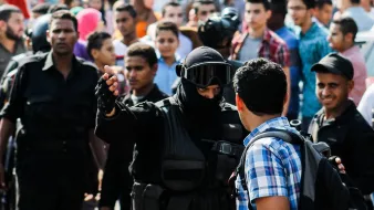 L'image montre une scène de confrontation dans un contexte urbain. À l'avant, un homme vêtu d'un uniforme noir, portant un casque et une visière, semble interagir avec un autre homme qui porte une chemise à carreaux. Ce dernier semble questionné ou arrêté, tandis qu'un groupe de personnes les entoure, observant la situation. On peut percevoir une atmosphère tendue, avec des expressions variées sur les visages des spectateurs. La scène suggère un moment de tension ou de dispute, probablement lié à une manifestation ou à une intervention policière.