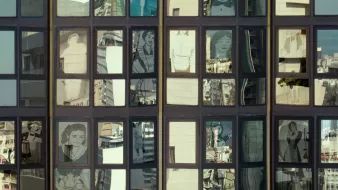 L'image montre une façade de bâtiment avec de grandes fenêtres. À travers ces fenêtres, on peut voir des reflets de la ville environnante ainsi que des illustrations ou des portraits de personnes qui semblent être intégrés dans les carreaux. Les illustrations sont stylisées et en noir et blanc, ajoutant un contraste intéressant avec les reflets colorés des bâtiments environnants. Cette composition crée une atmosphère visuelle dynamique et intrigante.