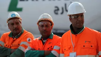 L'image montre trois ouvriers portant des vêtements de sécurité orange et des casques blancs. Ils se tiennent debout avec les bras croisés, affichant une attitude sérieuse. En arrière-plan, on peut voir un camion ou un véhicule avec le logo de Lafarge, une entreprise connue dans le secteur des matériaux de construction. L'environnement semble être un chantier ou une zone industrielle.