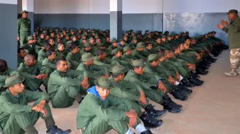 L'image montre un grand groupe de soldats assis dans une salle. Ils portent des uniformes verts et des bottes noires, et sont alignés en rangées sur le sol. Un instructeur, également en uniforme, se tient debout devant eux, semblant donner des instructions ou une formation. Les murs de la salle sont peints en gris, et l'atmosphère semble sérieuse et concentrée.