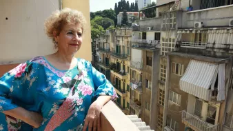 L'image montre une femme se tenant sur un balcon, avec une expression pensive. Elle porte une robe colorée ornée de fleurs. En arrière-plan, on peut voir un paysage urbain avec des bâtiments aux façades variées, certains anciennement entretenus et d'autres avec des voilages aux fenêtres. La lumière du jour illumine la scène, créant une atmosphère de tranquillité.