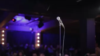 L'image montre un microphone sur un pied, au premier plan, sur une scène sombre. À l'arrière-plan, on aperçoit une audience floue et des lumières tamisées qui créent une ambiance de spectacle.