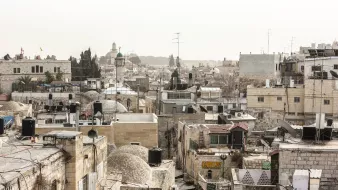 L'image montre un paysage urbain d'une ville ancienne, probablement Jérusalem. On peut voir une multitude de bâtiments en pierre avec des toits en tôle et des dômes, créant une texture variée. Les structures sont serrées les unes contre les autres, et quelques antennes de télévision sont visibles. Au loin, se distingue un minaret ou une coupole, indiquant la présence d'une mosquée. L'atmosphère semble un peu brumeuse, donnant une impression de chaleur et d'ancienneté à la scène.