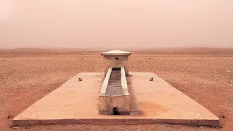 L'image montre une structure isolée située dans un paysage désertique. Au centre, il y a un long bassin en pierre, entouré par une surface plane et aride. Le sol est de couleur beige et semble sec et craquelé, tandis que l'atmosphère est légèrement brumeuse, ce qui donne une impression de chaleur et d'immensité désertique. Au loin, on aperçoit des collines ou des dunes qui se fondent avec le ciel dans une teinte douce et uniforme. L'endroit dégage une sensation de tranquillité et de solitude.